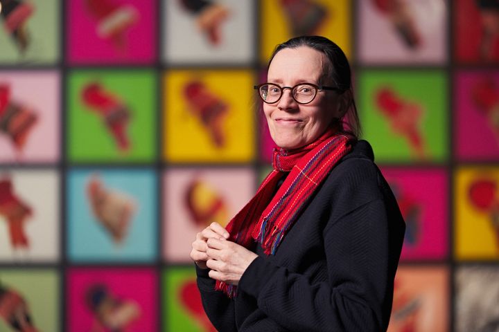 Sigga-Marja Magga seisoo värikkään taideteoksen edessä, katsoo hymyillen kameraan ja pitää punaista huivia.