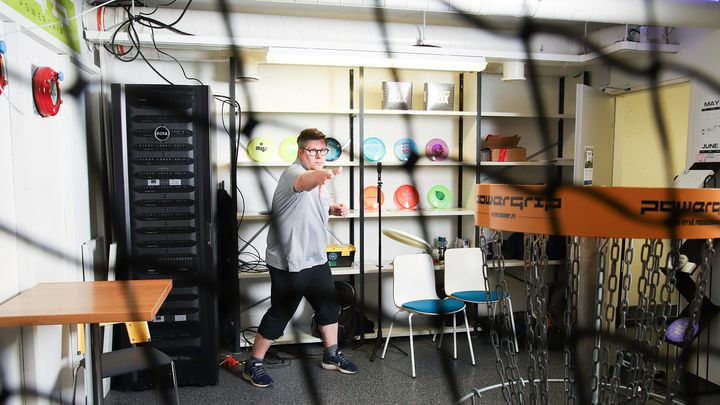 Kuvassa väitöskirjatutkija Marko Mustonen heittää frisbeetä sisätiloissa laboratoriossaan, jossa on frisbeegolfkori ja seinällä frisbeekiekkoja.