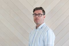 A man wearing glasses and a striped shirt stands against a wood-paneled wall.