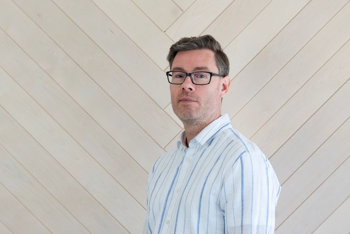 A%20man%20wearing%20glasses%20and%20a%20striped%20shirt%20stands%20against%20a%20wood-paneled%20wall.