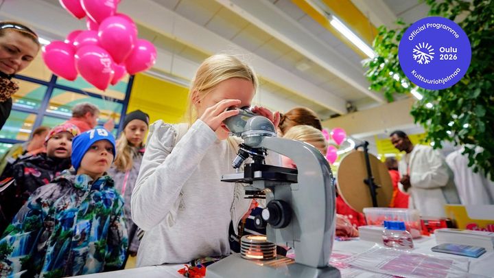 Lapsi katsoo mikroskooppiin Tutkijoiden yössä, taustalla on lapsia ja pinkkejä ilmapalloja.