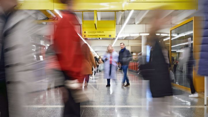 Eri-ikäisiä ihmisiä kulkee julkisen tilan käytävillä, osa kuvassa blurrattuina.