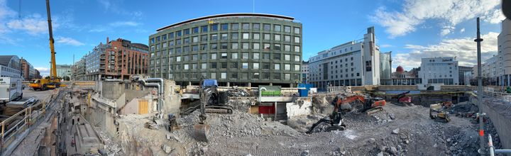 Työmaan laajakulmakuva, jossa purettua lippuhallirakennusta työkoneita ja monttu. Taustalla Kampin useita rakennuksia.