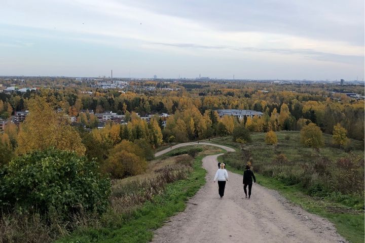 Kaksi henkilöä kävelee hiekkatietä alas Malminkartanonhuippua syksyisessä maisemassa, ympärillä oranssin ja keltaisen sävyisiä puita.