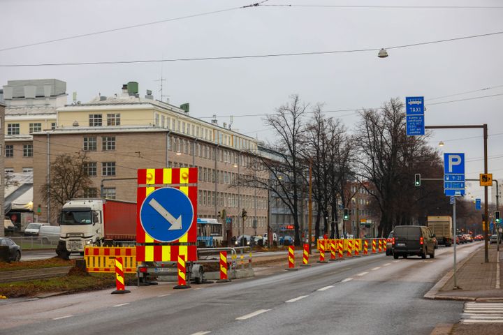Katuosuus, jossa on useita liikennemerkkejä ja kaistojen sulkemista osoittavia liikenne-esteitä. Taustalla rakennuksia ja pysäköintialueen merkkejä.