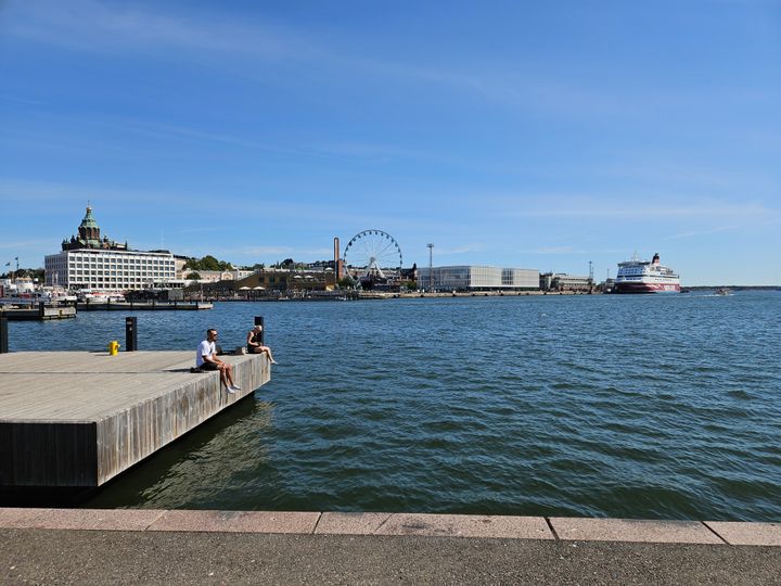 Ihmisiä merimaisemassa, taustalla matkustajalaiva satamassa.