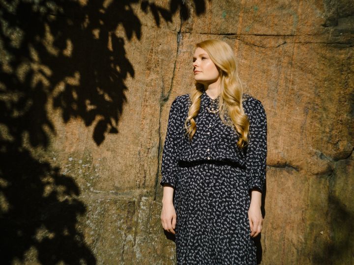 Kirjailija Saara Turunen seisoo ulkona auringonvalossa kiviseinän edessä.