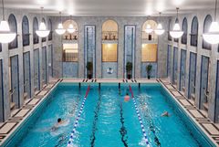 En blåskimrande simhallsvy öppnar sig mellan nyklacissicistika pelare som sträcker sig i två våningar. Simbassängen glittrar och det simmar några personer i den. Vackra lampor syns i den övre våningen.