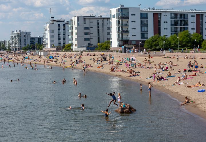 En sandstrand med många människor som badar och solar, samt moderna byggnader i bakgrunden.