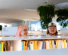 En kvinna och en man ser på varandra och ler, mellan bibliotekets bokhyllor.