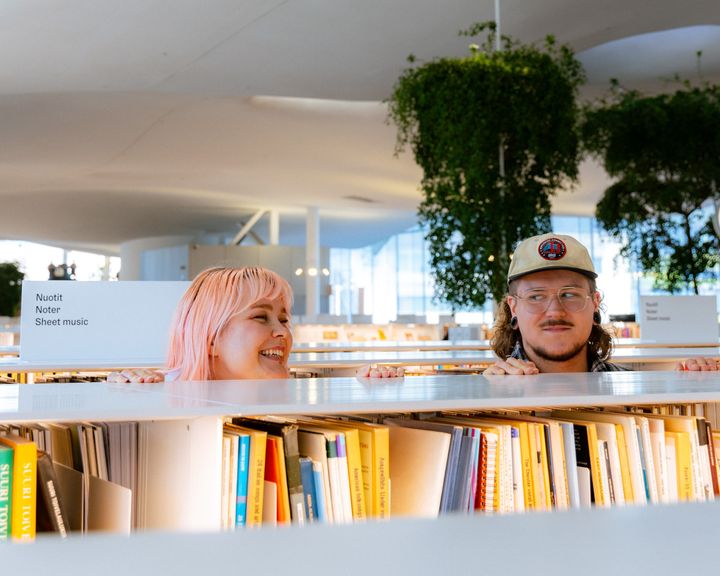 En kvinna och en man ser på varandra och ler, mellan bibliotekets bokhyllor.