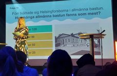 Kvinna står på scen framför en bild med frågetext om bastur samt en bild av ett gammalt hus. Flera svarsalternativ visas.