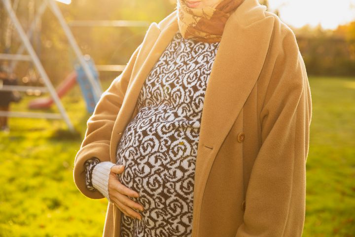 Gravid kvinna i beige kappa och mönstrad klänning håller handen på magen, stående utomhus vid en lekplats i gräs.