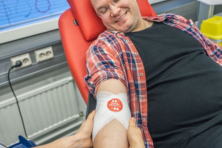 Mies istuu ja hymyilee verta luovutettuaan. Hoitaja on tehnyt kyynärtaipeeseen siteen, jossa on punainen Hengenpelastaja-tarra.