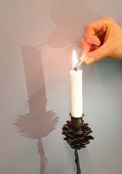 Käsi sytyttämässä kävyn mallisessa kynttilänjalassa olevaa valkoista kynttilää, jonka varjo heijastuu seinälle