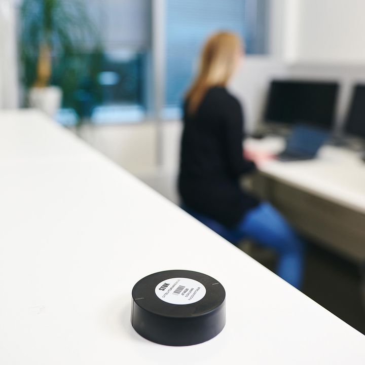 Radonpurkki hyllyllä työhuoneessa, jossa henkilö istuu työpöydän ja tietokoneen ääressä.