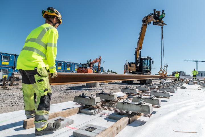 Aurinkoisena päivänä etualalla työntekijä ohjaa raitiotiekiskoa paikoilleen, kaivinkone nostaa kiskoa keskeltä ja taustalla toinen työntekijä ohjaa kiskoa paikoilleen.