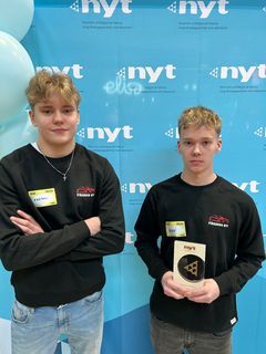 Kaksi nuorta seisoo kuvausseinäkkeen edessä Uskalla Yrittää -palkinto kädessä.