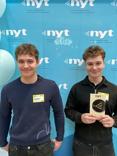 Kaksi nuorta seisoo kuvausseinäkkeen edessä Uskalla Yrittää -palkinto kädessä.