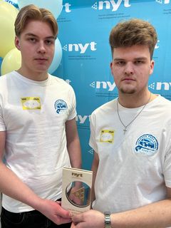 Kaksi nuorta seisoo kuvausseinäkkeen edessä Uskalla Yrittää -palkinnon kanssa.
