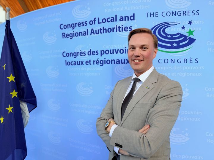 Jani Kokko seisoo hymyillen Euroopan neuvoston kunta- ja aluehallintokongressin kuvausseinän edessä.