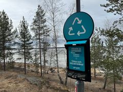 Skylt med återvinningssymbol och text om plogging i parkmiljö, omgiven av träd och sjöutsikt i bakgrunden.