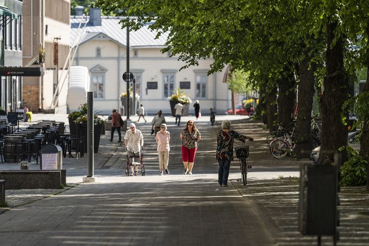 En stadsgata med människor som promenerar och cyklar i solskenet, omgivna av träd och byggnader.