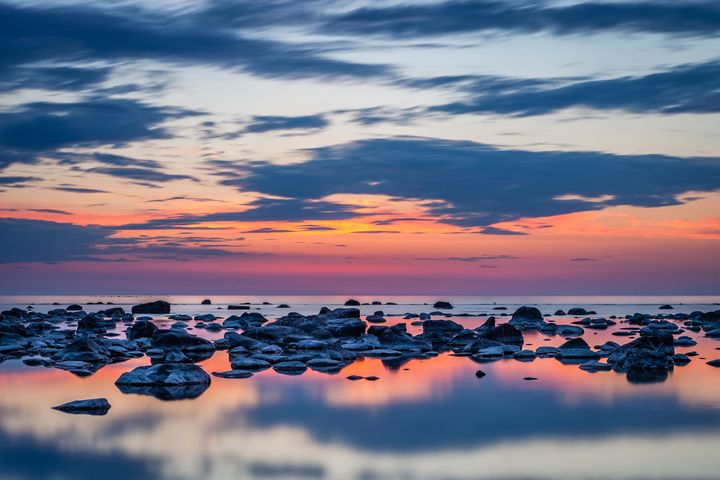 Auringonlasku kivikkoisella rannalla, taivas hehkuu oranssin ja sinisen sävyissä, heijastuksia vedessä.