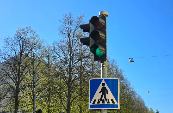 Liikennevalot, joissa palaa vihreä valo. Alla suojatien merkki. Taustalla puita ja sininen taivas.