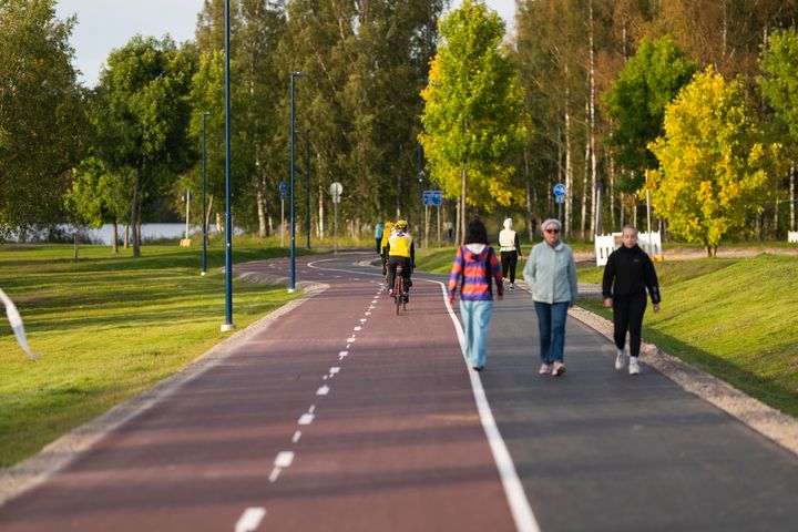 En cykel- och gångväg med flera personer som promenerar och en person som cyklar, omgiven av träd och grön gräsmatta.