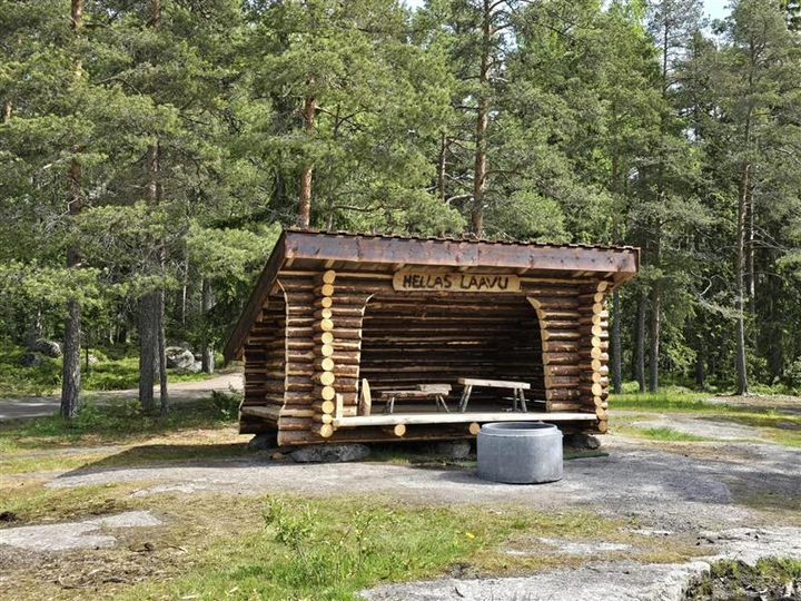Ett vindskydd byggt av timmer i skogsmiljö. Inuti vindskyddet finns två träbänkar. Ovanför ingången sitter en skylt med texten "Hellas Laavu".