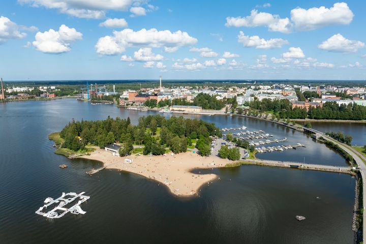 Flygbild av en stad med en sjö, strand, grönska och båtar vid en småbåtshamn.