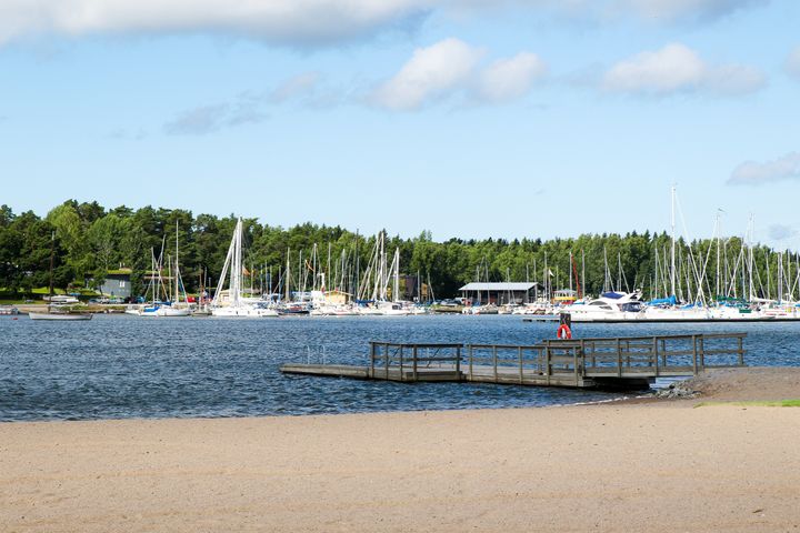 Brygga vid en sandstrand med segelbåtar i bakgrunden och träd längs horisonten under en blå himmel.