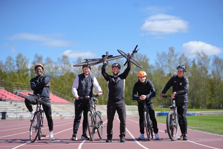 Fem personer poserar med cyklar på en löparbana under en solig dag.