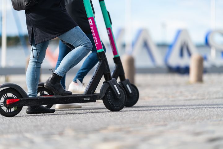 Två personer åker elsparkcyklar på en stenbelagd väg nära vattnet.