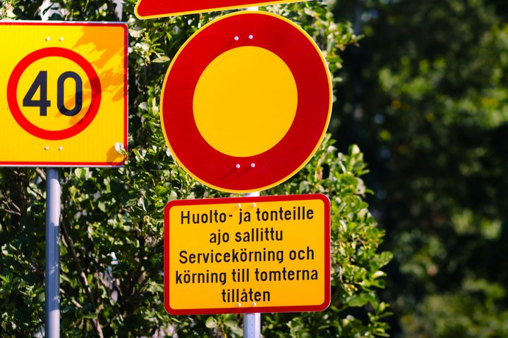 Trafikskyltar mot gröna buskar, inklusive en skylt med text på finska och svenska om tillåten servicekörning.