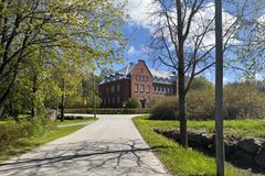 Stort rött tegelhus med vita fönster omgiven av grönska under en solig himmel. En asfalterad väg leder fram till det.