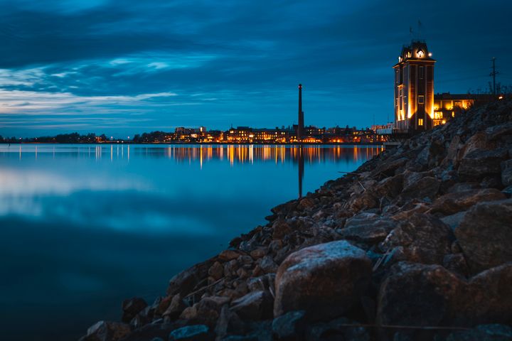 Merimaisema iltahämärässä, rannalla kiviä, taustalla valaistuja rakennuksia.