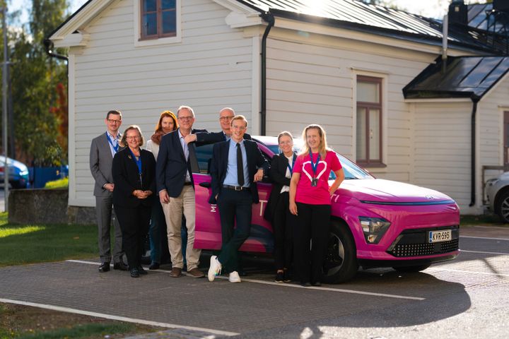 A group of Vaasa's senior city officials standing and smiling beside a pink electric car in front of a wooden building.