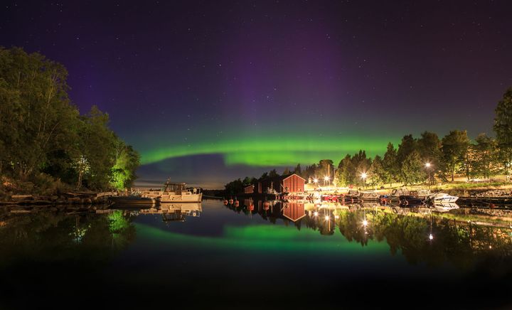Norrskenet lyser i grönt och violett. Ett strandlandskap där vattnet speglar färgerna. I bakgrunden syns röda stugor och träd.