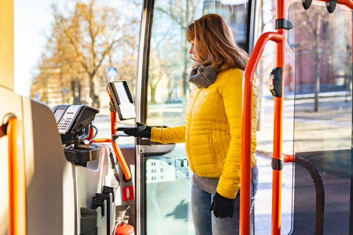 Nainen nousee bussiin kirkkaana päivänä, pukeutuneena keltaiseen takkiin ja harmaaseen huiviin.