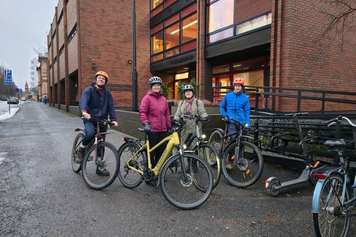 Fyra personer med cyklar står på en asfalterad väg framför en tegelbyggnad. De har cykelhjäl