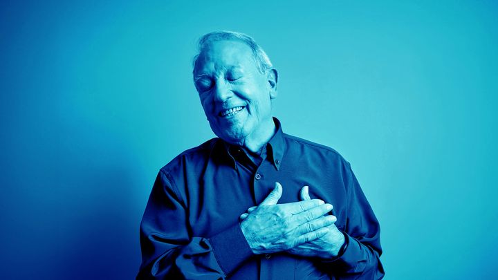 Vanhempi mies, jolla on kädet sydämen päällä ja joka hymyilee sinisessä valossa.