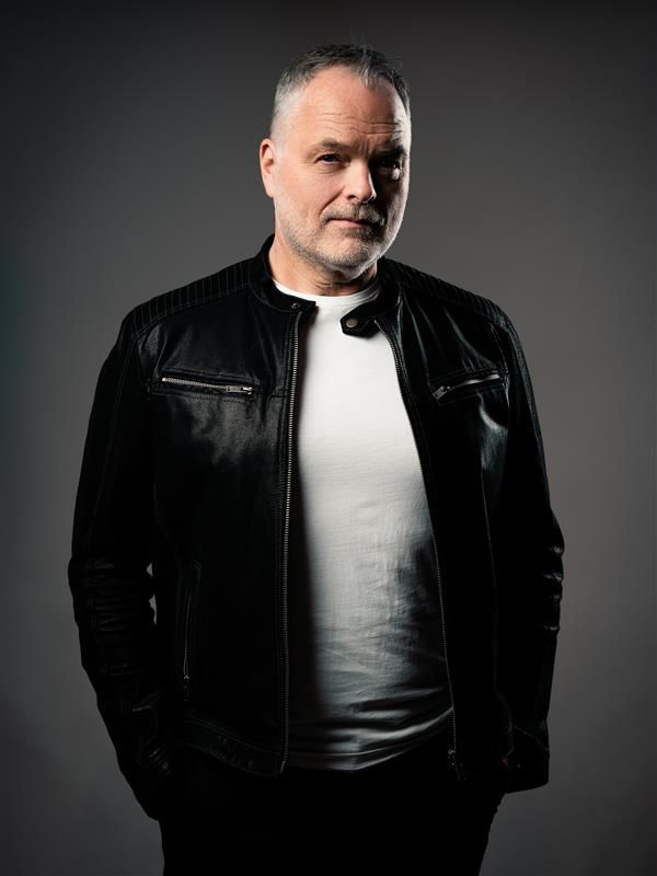 En man står framför en grå bakgrund klädd i en läderjacka och en vit T-shirt och tittar in i kameran.