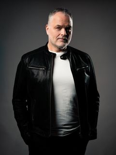 En man står framför en grå bakgrund klädd i en läderjacka och en vit T-shirt och tittar in i kameran.