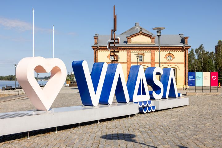 Teksti "VAASA" isoilla kirjaimilla ulkona, sydämen muotoinen symboli vasemmalla. Taustalla punatiilinen rakennus ja sininen taivas.