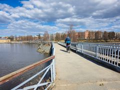 En person cyklar över en bro med vatten på ena sidan under en molnig himmel.