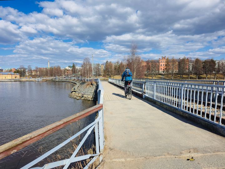 En person cyklar över en bro med vatten på ena sidan under en molnig himmel.