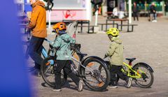 Två barn i hjälmar leder sina cyklar bredvid en vuxen person på ett torg.