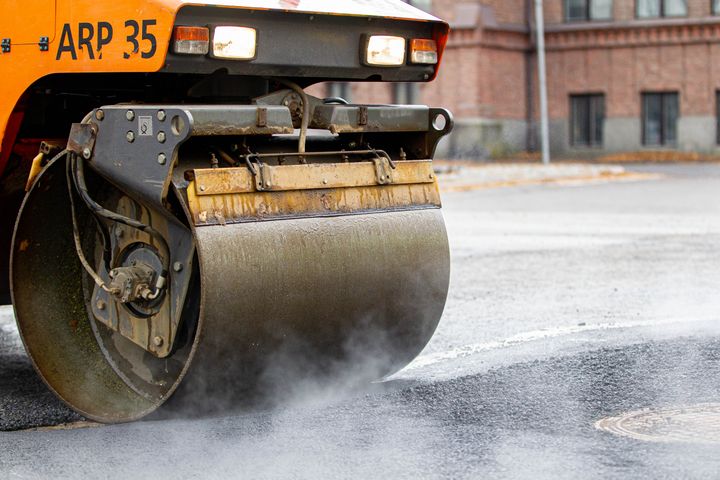 Tiehöylä tasoittaa asfaltoitua tietä. Taustalla näkyy tiilirakennus.